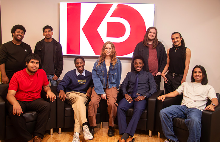 Spring 2026 acting graduates, some sitting in chairs some standing behind them, with the KD Studio logo on a large TV screen behind them.